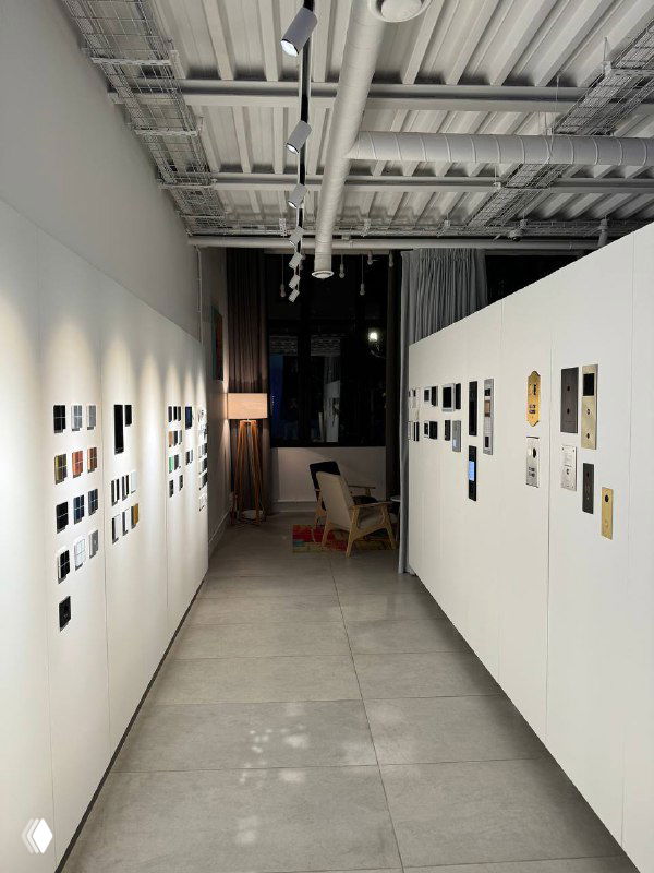 Коридор небольшой выставочной галереи с белыми стендами и развешенными работами; в глубине видны кресло и тёмное окно.