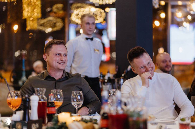 Гости за праздничным столом в панорамном ресторане: улыбающиеся мужчины, бокалы и блюда на переднем плане, праздничные огни