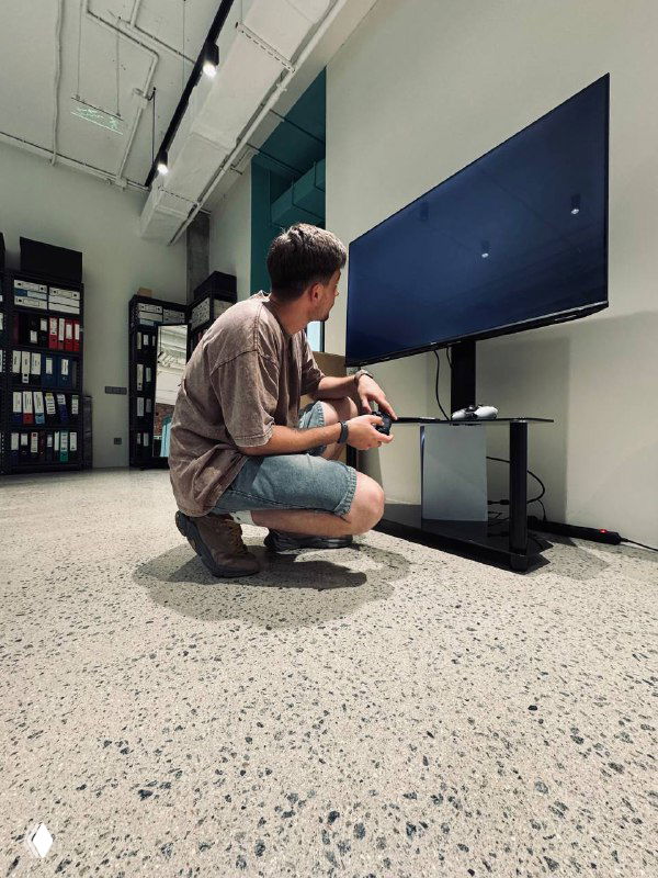 Мужчина в магазине электроники присел перед большим телевизором, держит пульт и проверяет его подключение и экран.