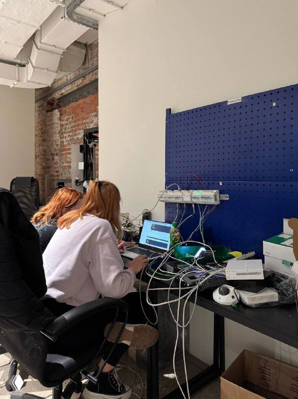 Два сотрудника сидят за рабочим столом в офисе и работают за ноутбуками; на столе техника, провода и другие предметы офисной среды, не связанные с едой.