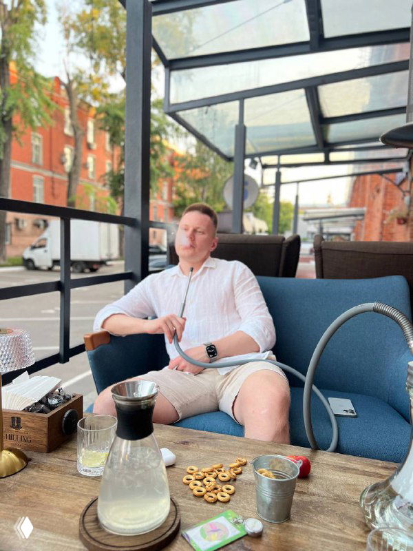 Мужчина в светлой рубашке и шортах сидит в уличном кафе на диване за столиком с кальяном и напитками; на заднем плане кирпичные дома и проезжая часть.