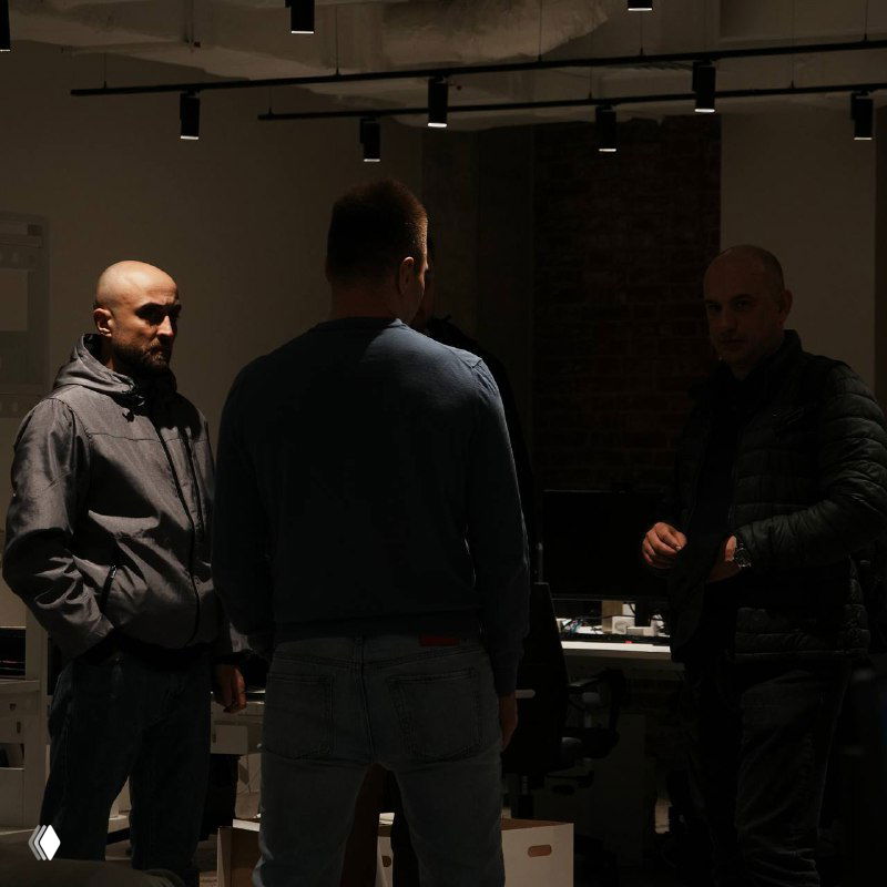 Три мужчины в офисе при приглушённом свете обсуждают рабочие вопросы; снимок подчёркивает деловой контекст.