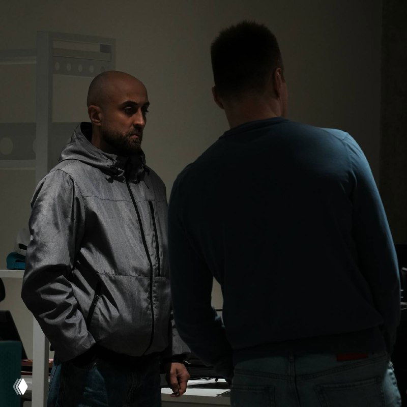 Два мужчины в офисе при тусклом освещении ведут беседу у рабочих мест; кадр подчёркивает деловую атмосферу.