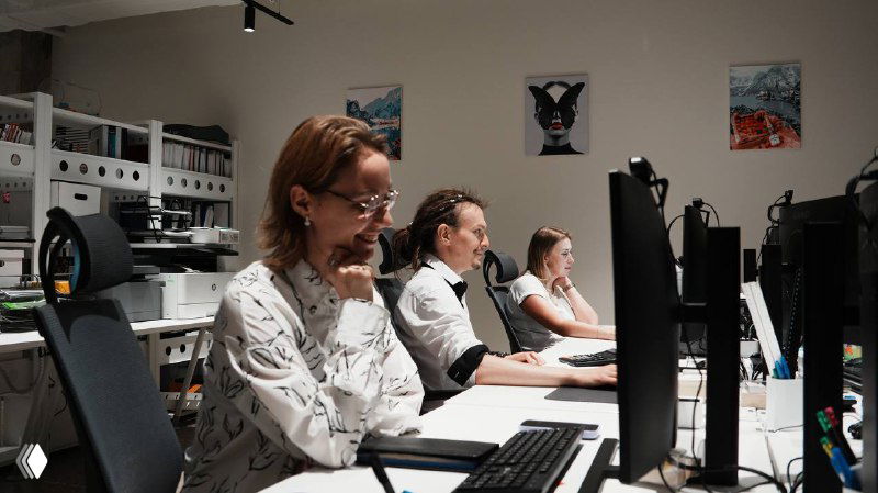 Фото офиса: несколько сотрудников за мониторами в открытом рабочем пространстве, люди работают вместе в атмосфере корпоративной команды.