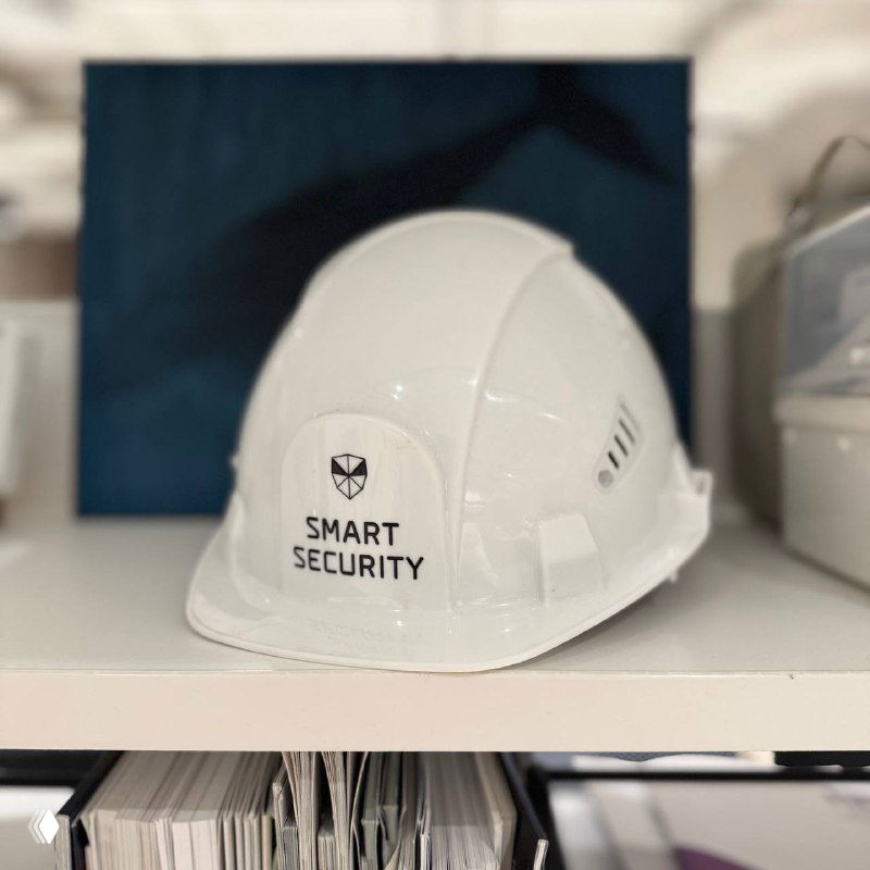 Белая защитная каска с надписью «SMART SECURITY» на полке; нейтральная фотография, иллюстрирующая тему найма и профессиональной ответственности.