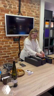 Сцена упаковки: женщина за столом завязывает ленту на подарочной коробке, подготовка корпоративных наборов в офисе.