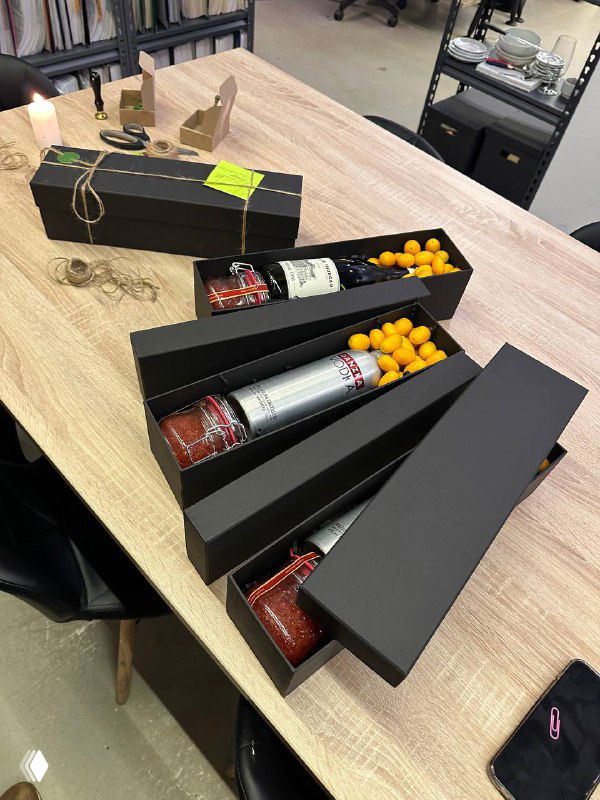 Три открытых черных коробки с бутылками алкоголя, желтыми мандаринами и упаковками деловых подарков на деревянном столе.