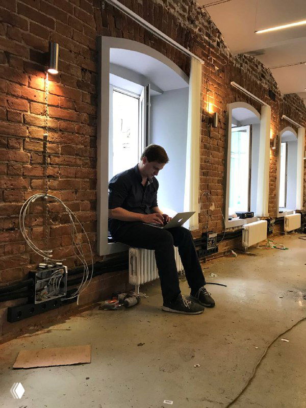 Мужчина с ноутбуком сидит на радиаторе у высокой кирпичной стены в помещении на ремонте — рабочая зона в процессе обустройства и подготовки офиса.