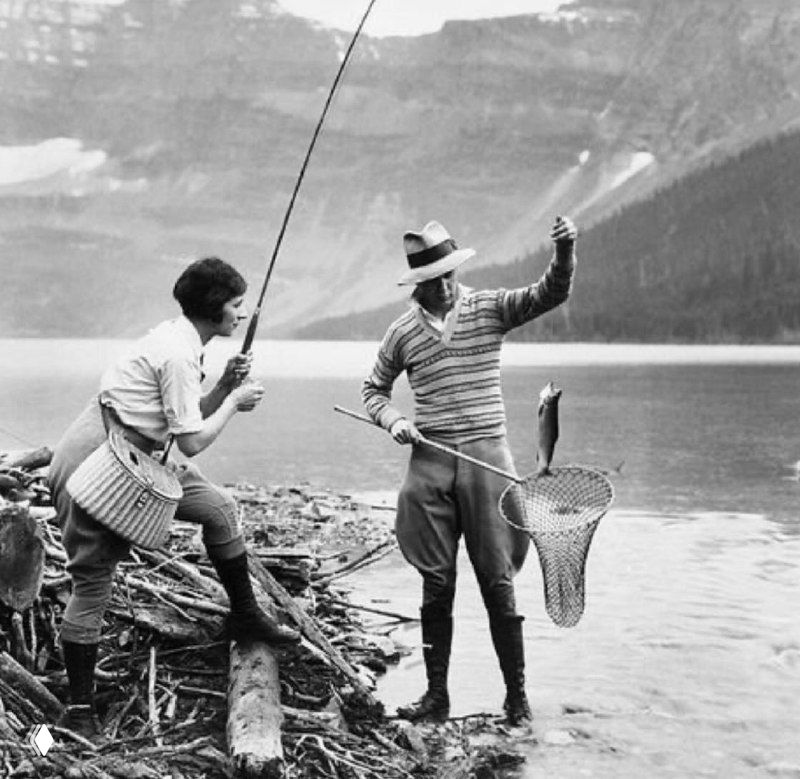 Черно‑белая фотография двух рыбаков у озера: один держит удочку, другой поднимает рыбу в подсачеке на фоне гор и спокойной воды, винтажная сцена.