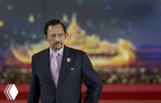 Портрет султана Хассанала Болкиаха в официальной обстановке; мужчина в костюме на фоне сценического оформления и подсветки, формальный ракурс.