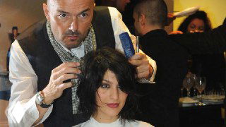 Парикмахер работает с клиенткой в салоне: видны руки мастера, ножницы и интерьер заведения с посетителями на фоне, бытовая сцена.