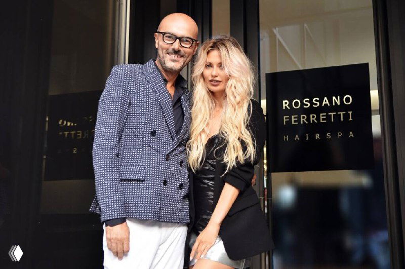 Россано Ферретти рядом с женщиной у входа в салон Rossano Ferretti Hairspa; видна вывеска бренда и фасад салона, деловая и брендовая фотография.