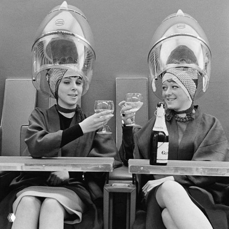 Две клиентки в салоне под сушуарами поднимают бокалы и держат бутылку шампанского — атмосфера летней акции и дружелюбного сервиса.