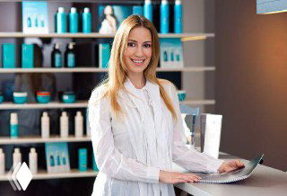 Улыбающийся сотрудник салона у стойки регистрации на фоне полок с профессиональной косметикой, рабочая сцена приёма клиентов.