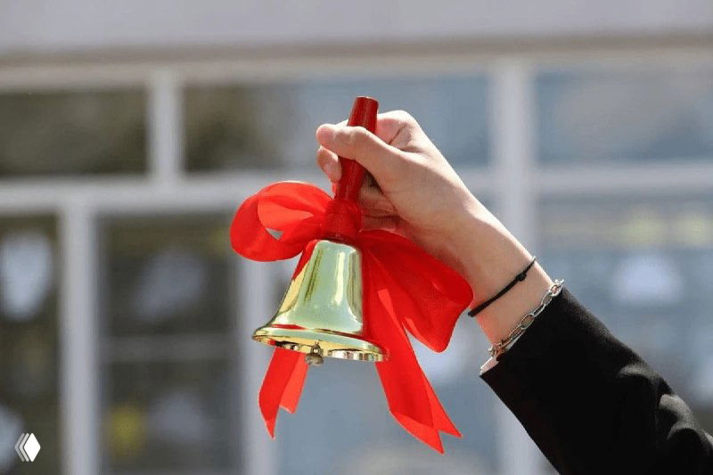 Рука держит церемониальный колокольчик с красной лентой на размытом фоне здания — символ старта обучения и делового события.