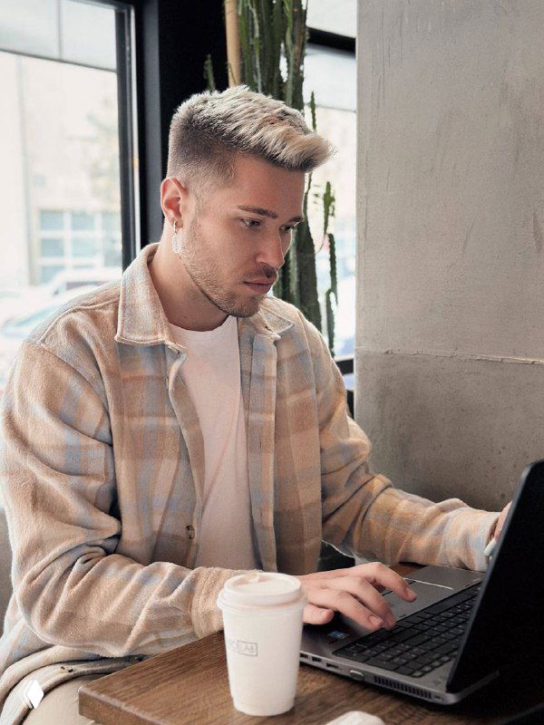 Человек сидит в кафе за ноутбуком и держит бумажный стаканчик кофе, деловой кадр для иллюстрации интервью и работы в beauty-маркетинге.