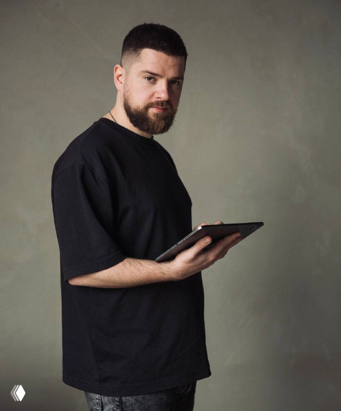 Портрет Максима Спивака в студии: мужчина с бородой в тёмной футболке держит планшет на фоне нейтральной стены, деловой образ для интервью.