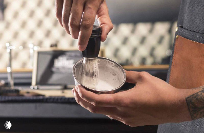 Барбер взбивает пену в металлической чаше, видны руки мастера и рабочая поверхность салона — иллюстрация мужского премиального ухода и услуги.