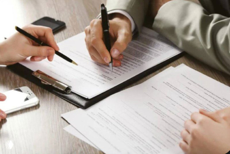 Руки двух людей подписывают документы на столе: бланки, ручки и смартфон, что иллюстрирует оформление регламента и служебных правил.