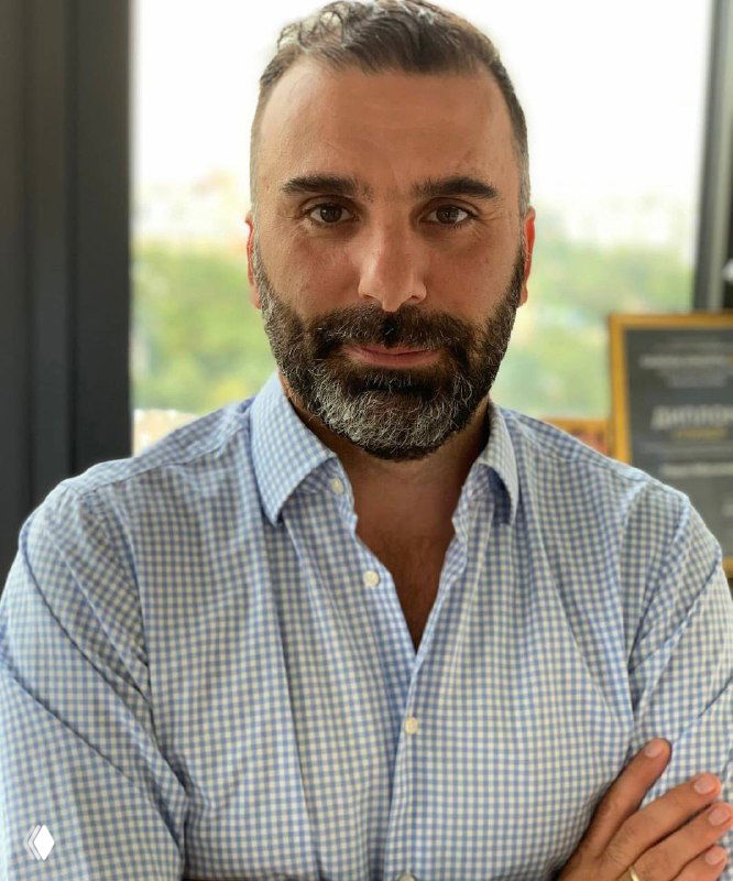 Портрет мужчины в светлой клетчатой рубашке с бородой и легкой щетиной на фоне окна; иллюстрация интервью о создании сервиса KPI.bi и развитии аналитики для бизнеса.