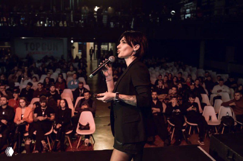 Женщина-спикер на сцене с микрофоном перед полной аудиторией; кадр с делового мероприятия бьюти-индустрии, интервью или лекция в зале.