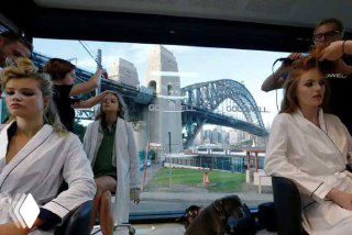 Модели готовятся в салоне красоты в Сиднее; через окно виден Харбор-Бридж и городской пейзаж — подготовка перед показом мод.