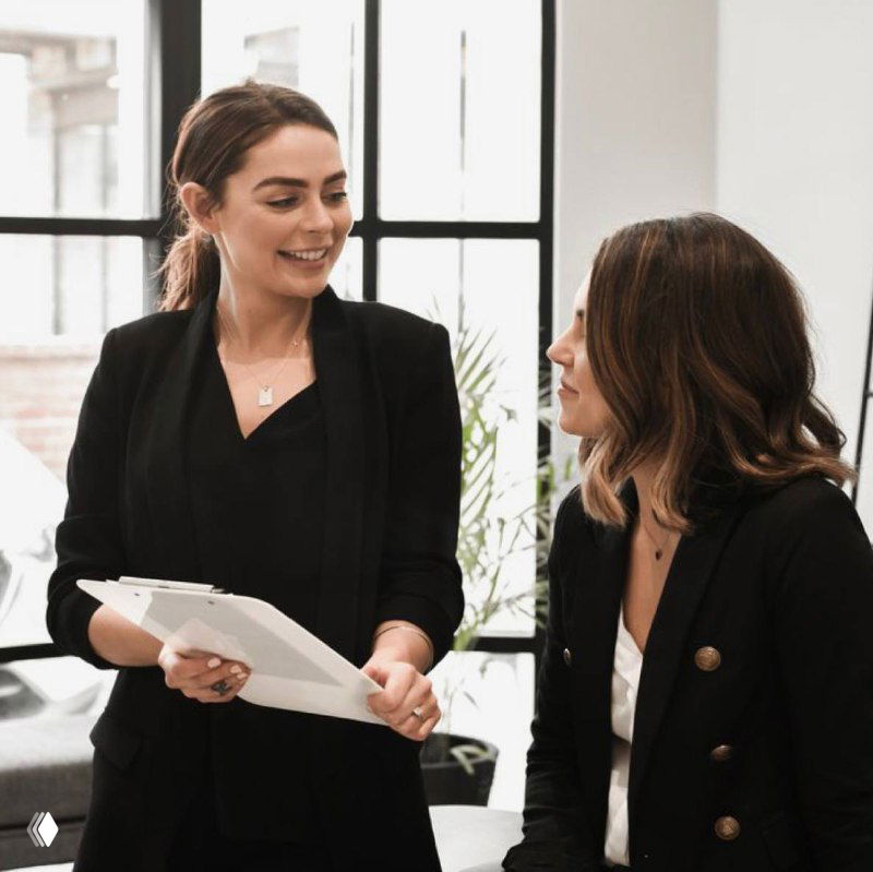 Две сотрудницы в офисе обсуждают индивидуальный план развития: менеджер держит папку с документами, ведётся деловой разговор у окна.