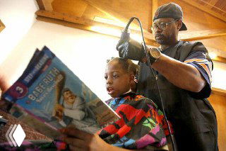 Парикмахер стрижет ребёнка, который читает книгу вслух; сцена иллюстрирует инициативу Кортни Холмс из Айовы и тёплую атмосферу сервиса.