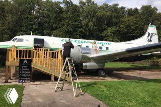 Внешний вид салона красоты, обустроенного в винтажном самолёте «Амбер»: самолёт с подъёмной лестницей и входной площадкой на фоне зелёных насаждений.