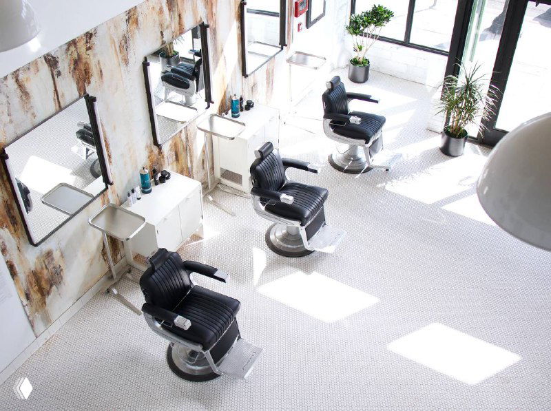Интерьер парикмахерской в ретро-стиле: ряд чёрных кресел для стрижки, зеркала вдоль стены и яркое естественное освещение в просторном помещении.