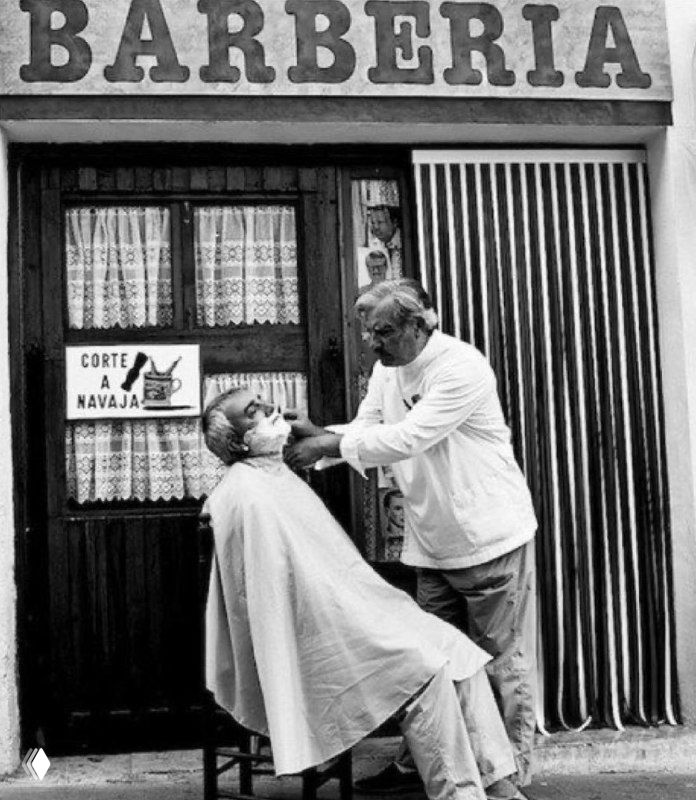 Черно-белое архивное фото: барбер стрижет клиента у входа в барбершоп с вывеской «BARBERIA», ретро-атмосфера уличного кадра.