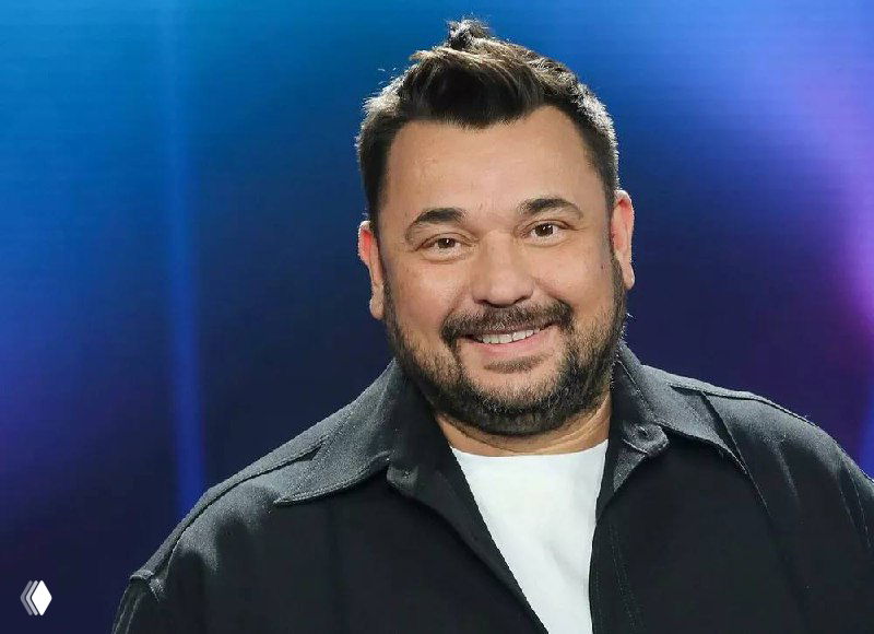 Портрет мужчины на сцене в тёмной одежде, улыбается на студийном фоне — иллюстрация к новости о запуске бьюти-бренда.