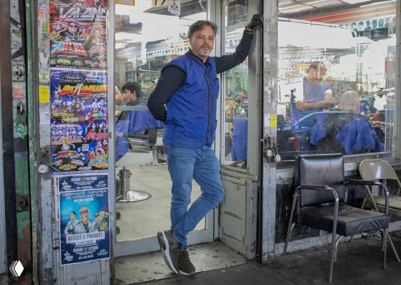 Барбер стоит в дверном проёме барбершопа на фоне кресел и вывесок — сцена у входа Langumas El Cache в Квинсе, Нью-Йорк, после инцидента