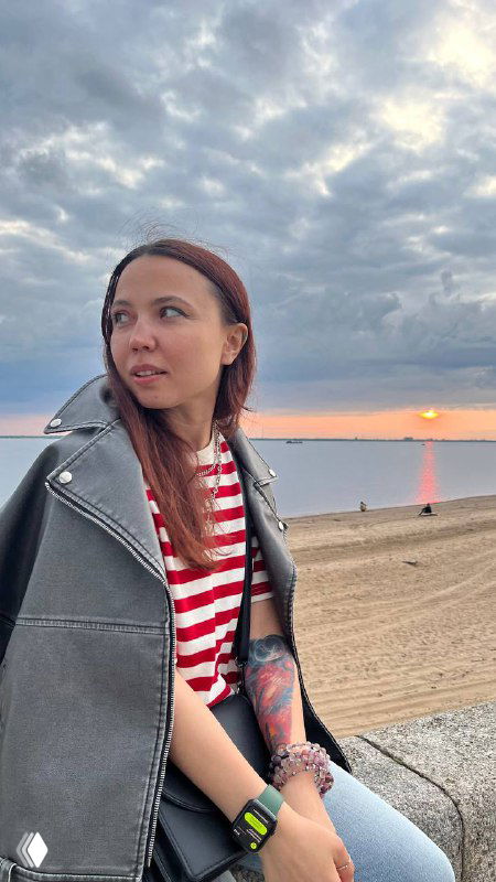 Женщина в полосатой футболке и кожаной куртке стоит на набережной у моря на фоне пасмурного неба и заката, задумчиво смотрит в сторону.