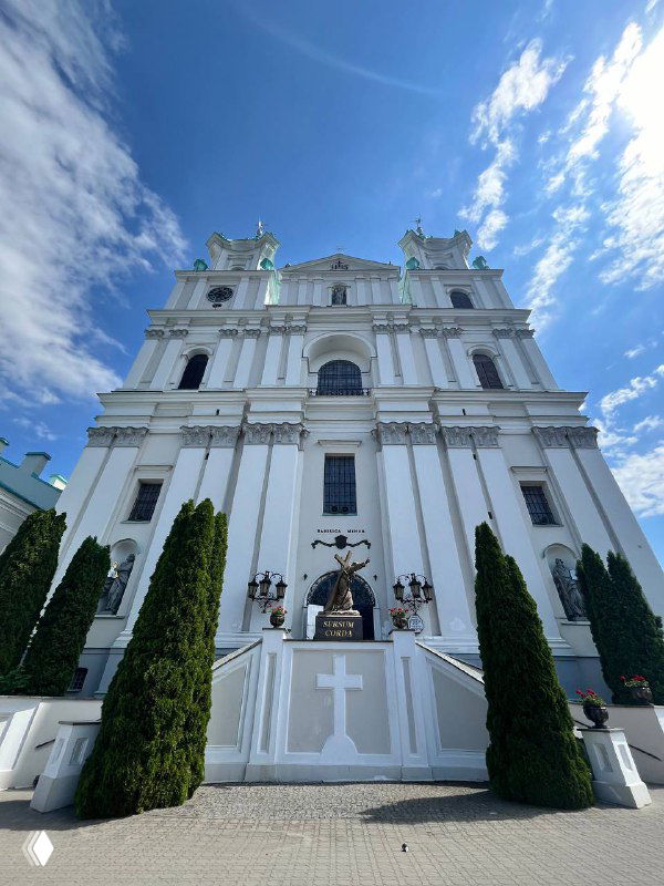 Величественный фасад костёла на фоне насыщенно-голубого неба с облаками; архитектурные детали, ступени и крест у входа.