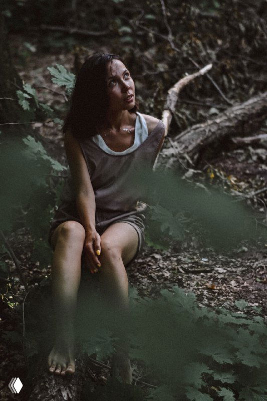 Девушка сидит на земле в лесу, взгляд в сторону, мягкий свет и размытый передний план создают ощущение интимного размышления и внутренней честности.