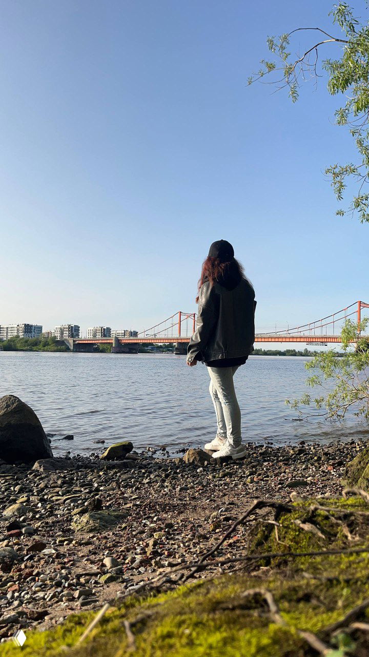 Женщина стоит на каменистом берегу реки, спиной к камере; на фоне виден мост и город, снимок передаёт задумчивое состояние и момент перемен в жизни.