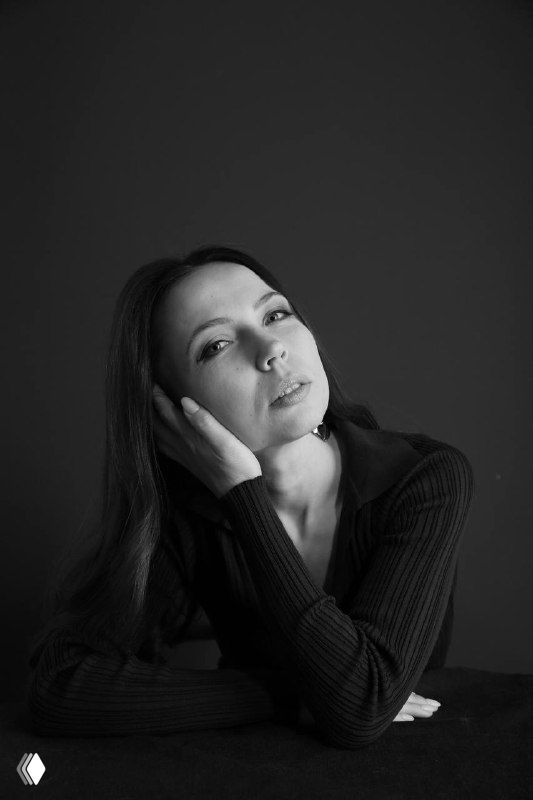 Черно-белый портрет женщины в задумчивой позе, опирающейся рукой на стол; тёмный фон, спокойный и рефлексивный образ автора поста.