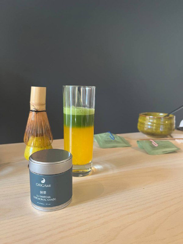 Высокий стакан с послойным зелёным и жёлтым напитком на столе, рядом банка с порошком матчи и бамбуковый венчик — акценты на напитках из матчи.