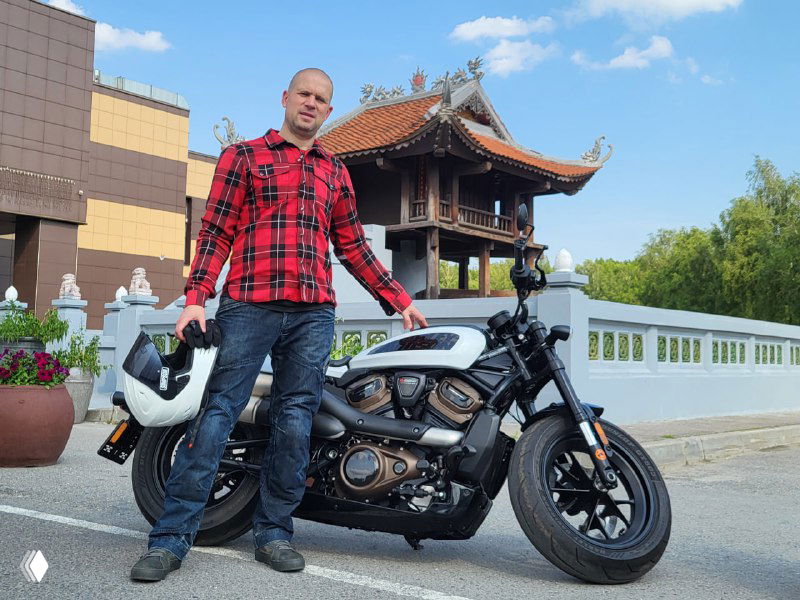 Николай рядом с Harley-Davidson Sportster S 1250 на стоянке у архитектурного павильона: мотоцикл в профиль, автор в клетчатой рубашке держит шлем.