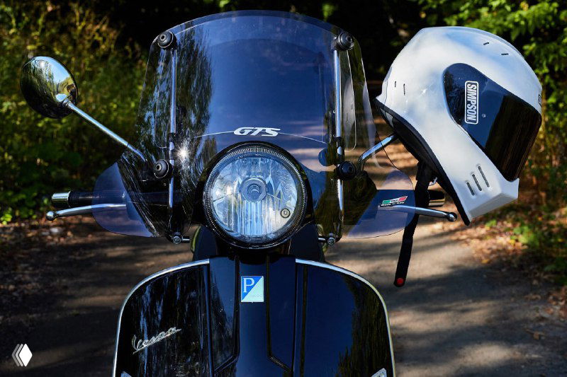 Чёрный скутер спереди с высоким ветровым стеклом и шлемом, надетым на зеркало, припаркованный на дороге среди зелёной растительности, символ мотосезона.