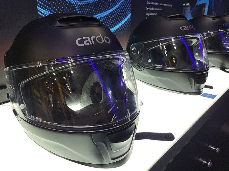 На переднем плане два матовых чёрных шлема Cardo Beyond с прозрачными визорами и логотипом Cardo — вид фронтально на выставочном стенде.