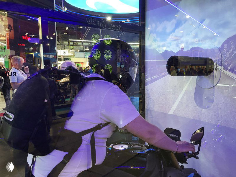 Посетитель выставки демонстрирует шлем/установку на стенде: человек в белой футболке пробует коммуникативный модуль Cardo на тренажёре на фоне экспозиции.