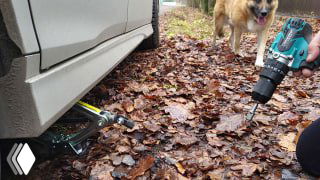 Рука с шуруповёртом и переходником вращает штатный домкрат под приподнятой машиной; вокруг осенняя листва, рядом собака.