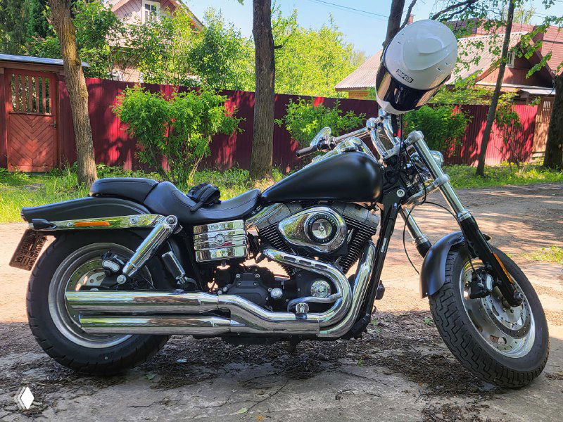 Черный Harley-Davidson, припаркованный во дворе рядом с красным забором; вид сбоку, видны хромированные глушители, двигатель и шлем.