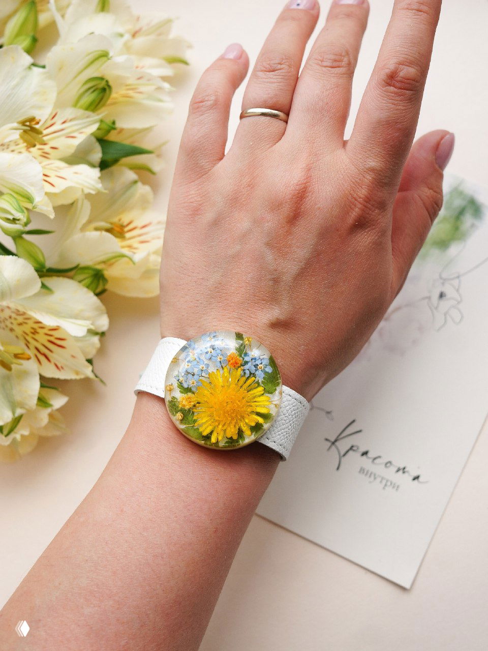 Крупный план браслета со смоляной круглой вставкой: внутри яркий жёлтый одуванчик и мелкие голубые незабудки на руке в белом ремешке