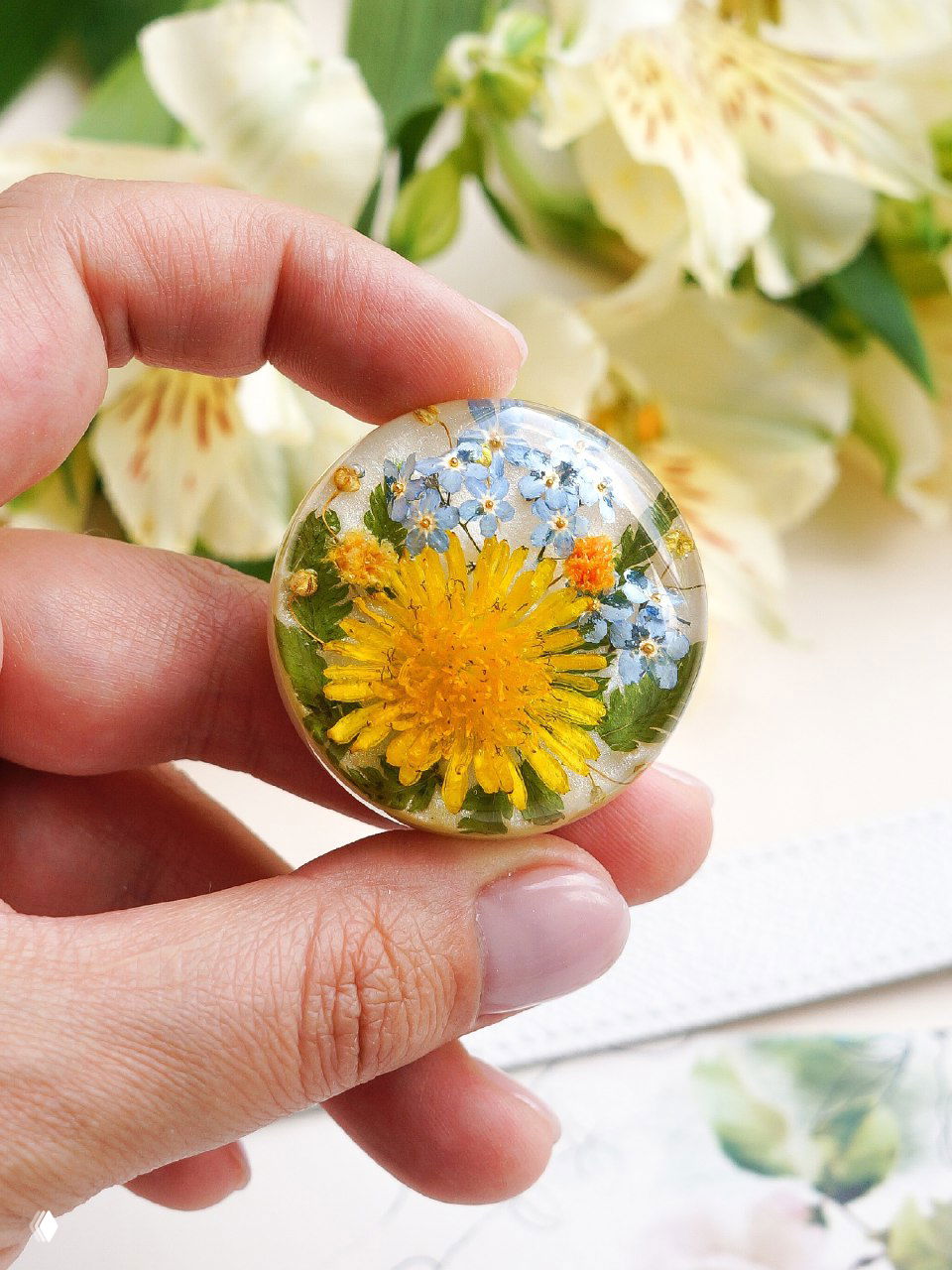 Фрагмент: автор держит круглую смоляную вставку с ярким жёлтым одуванчиком и мелкими синими цветочками, фон — цветы и белая бумага