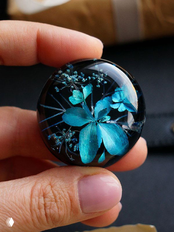 Круглая смолянная брошь с засушенными цветами голубых оттенков и тонкими стеблями зонтичного растения, держат пальцами.