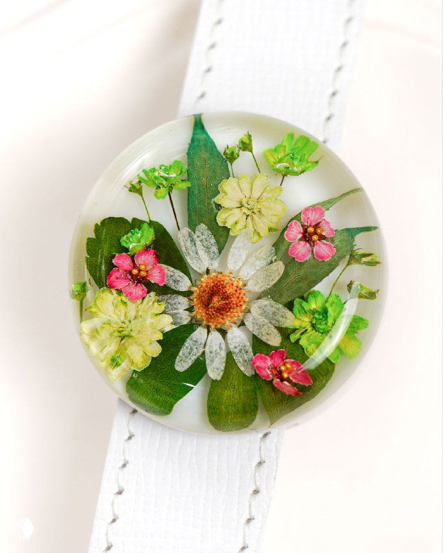 Крупный план круглого кабошона из прозрачной смолы с ромашкой, мелкими розовыми цветами и зелёными листочками на светлом фоне, макро.