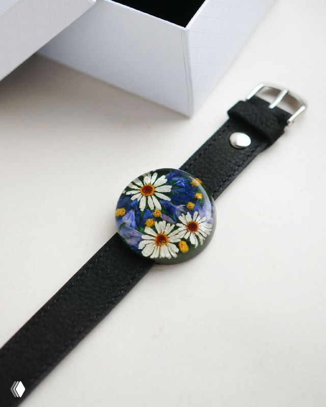 Чёрный кожаный ремешок с круглым смоляным украшением, внутри несколько белых ромашек на тёмно-синем фоне; минималистичная композиция.